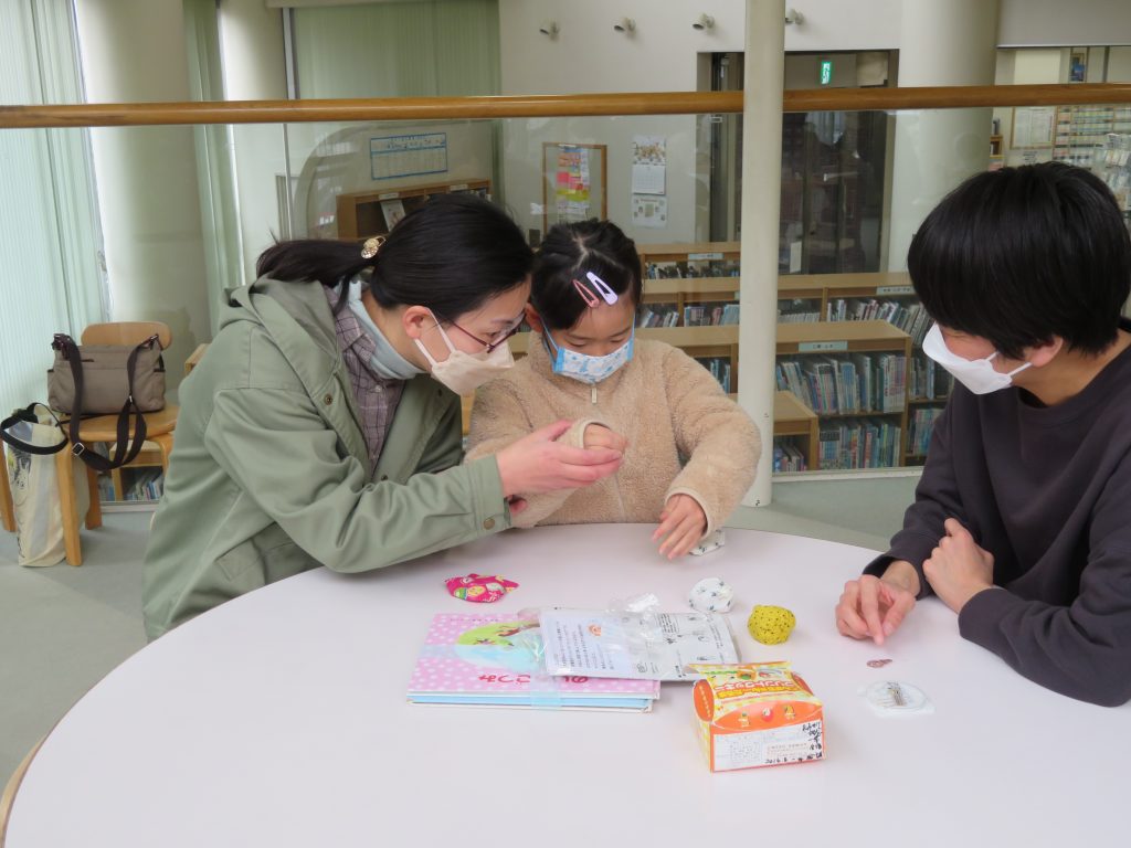 子供と二人の大人がテーブルで何かを作成している様子を描いた画像。