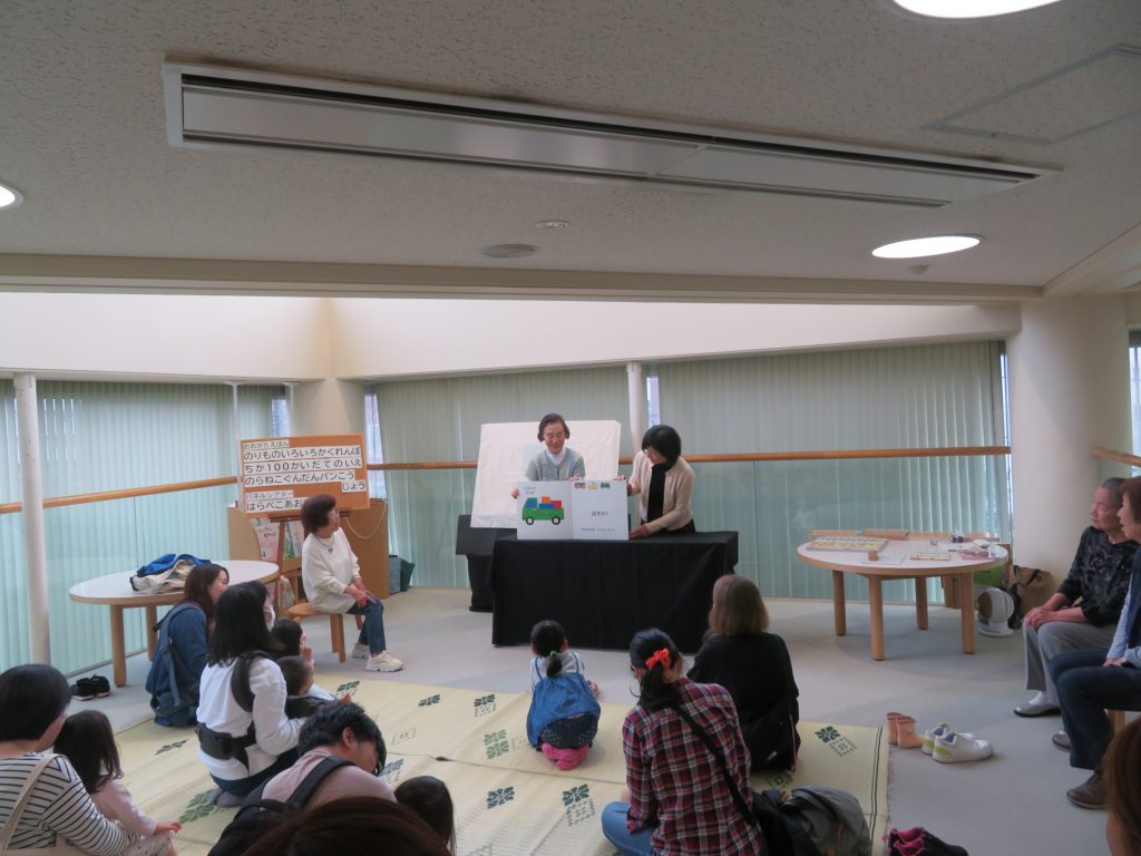 子供たちが集まる場所で、講師がテーブルの前で何かを説明している様子が描かれている。