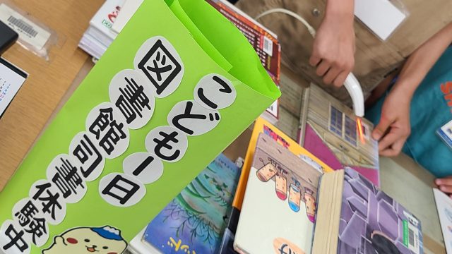 緑色の看板に「こども図書館日」という文字が書かれ、周囲には本が並んでいる様子。