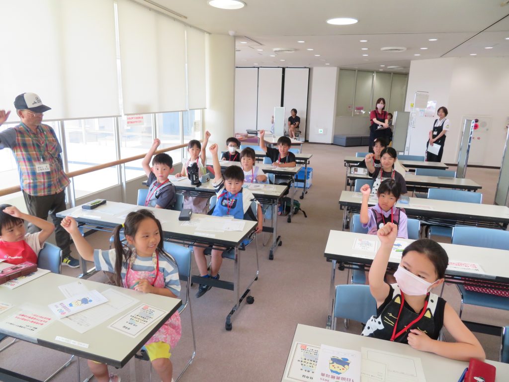 教室で手を挙げる子どもたちと大人のスタッフがいる風景。