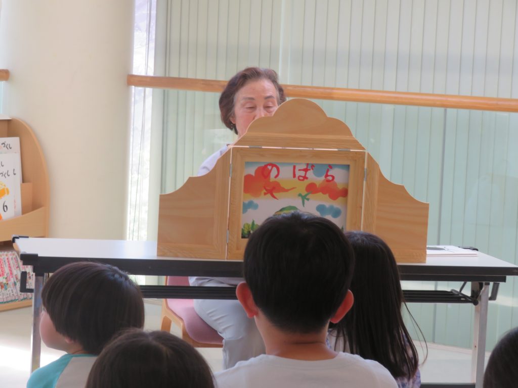 子供たちが物語を聞いている様子を写した画像で、前方には絵本が置かれたテーブルがある。