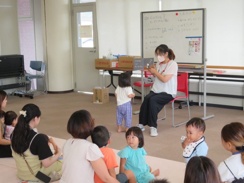 子供たちが座っている中で、椅子に座った大人が何かを見せている場面。