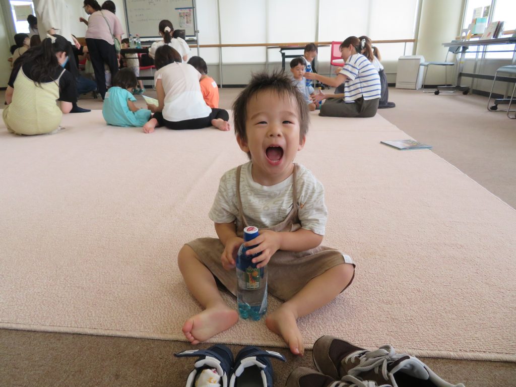 子供が座って飲み物を持ち、背景には他の人々が遊んでいる様子が見える部屋の写真。