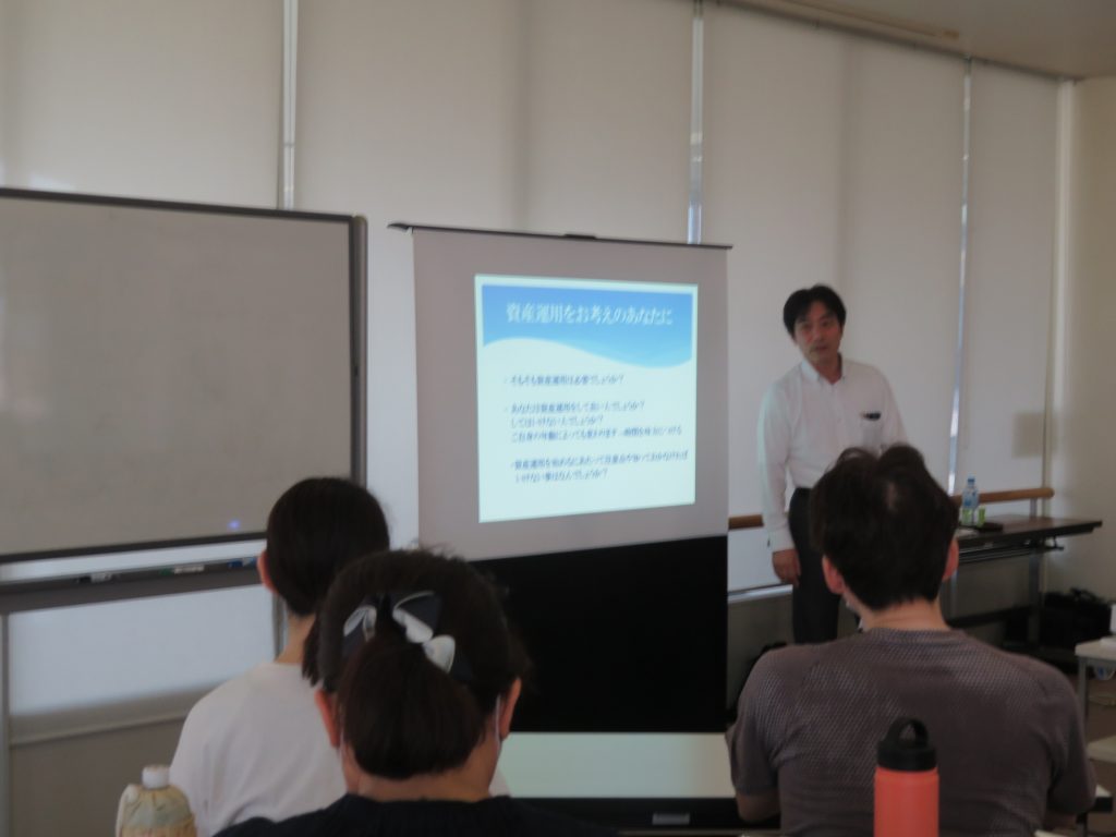 教室でプレゼンテーションを行っている人物とそれを聞いている受講者の後ろ姿が映っている画像です。