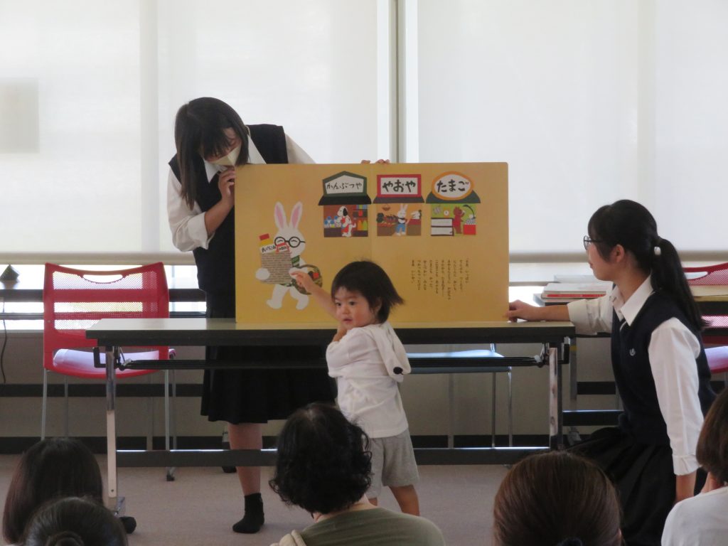 子供が絵本を見ながら話を聞いている様子を示す教室の風景。