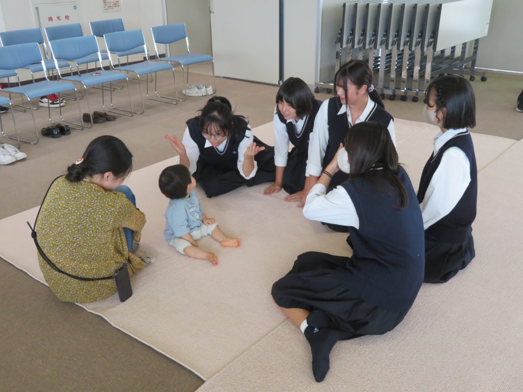 数人の大人が幼児と一緒に遊んでいる室内の様子。