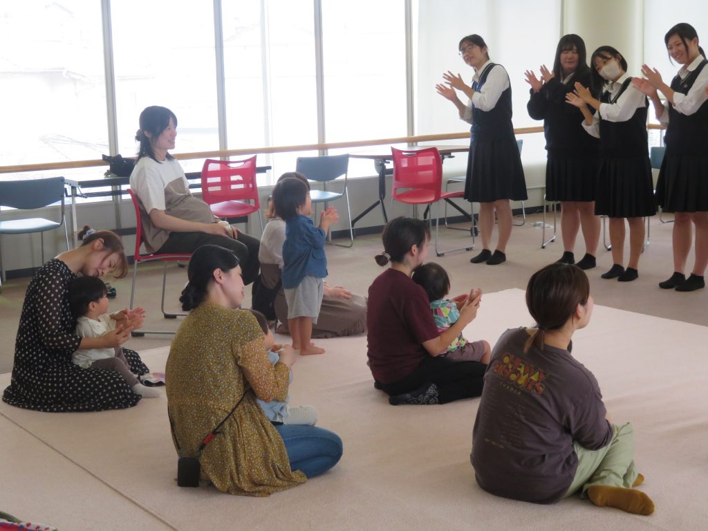 子供たちと大人たちが集まり、楽しそうに手を叩いている室内の様子。