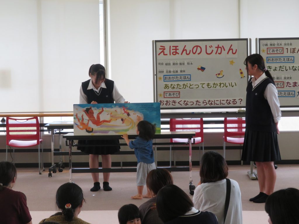 子供が絵本を持っている二人の大人に向かって話しかけている様子が描かれた教室の風景。