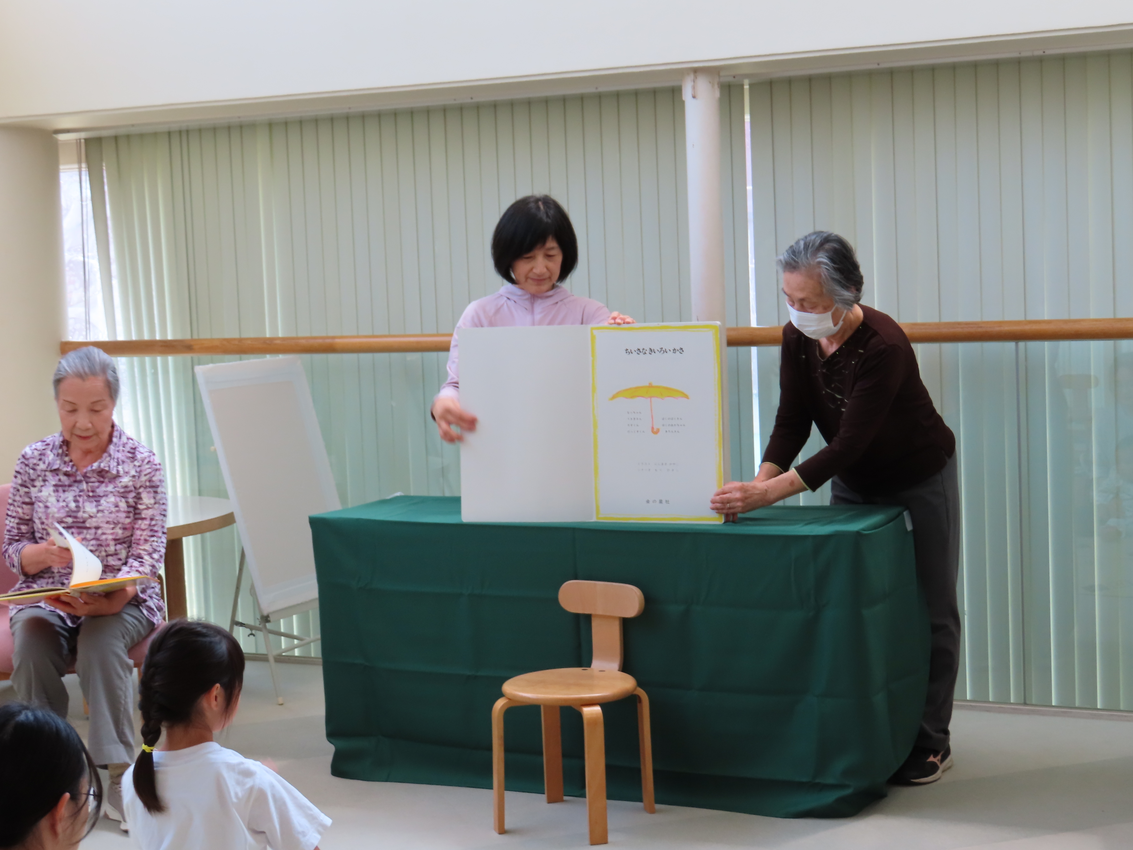 ボランティア二人がテーブルの上で大型の絵本を見せながら幼い子どもたちに読み聞かせをしている場面