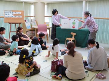 屋内で子どもと大人が床に座りボランティアがボードを使って読み聞かせや説明をしている様子
