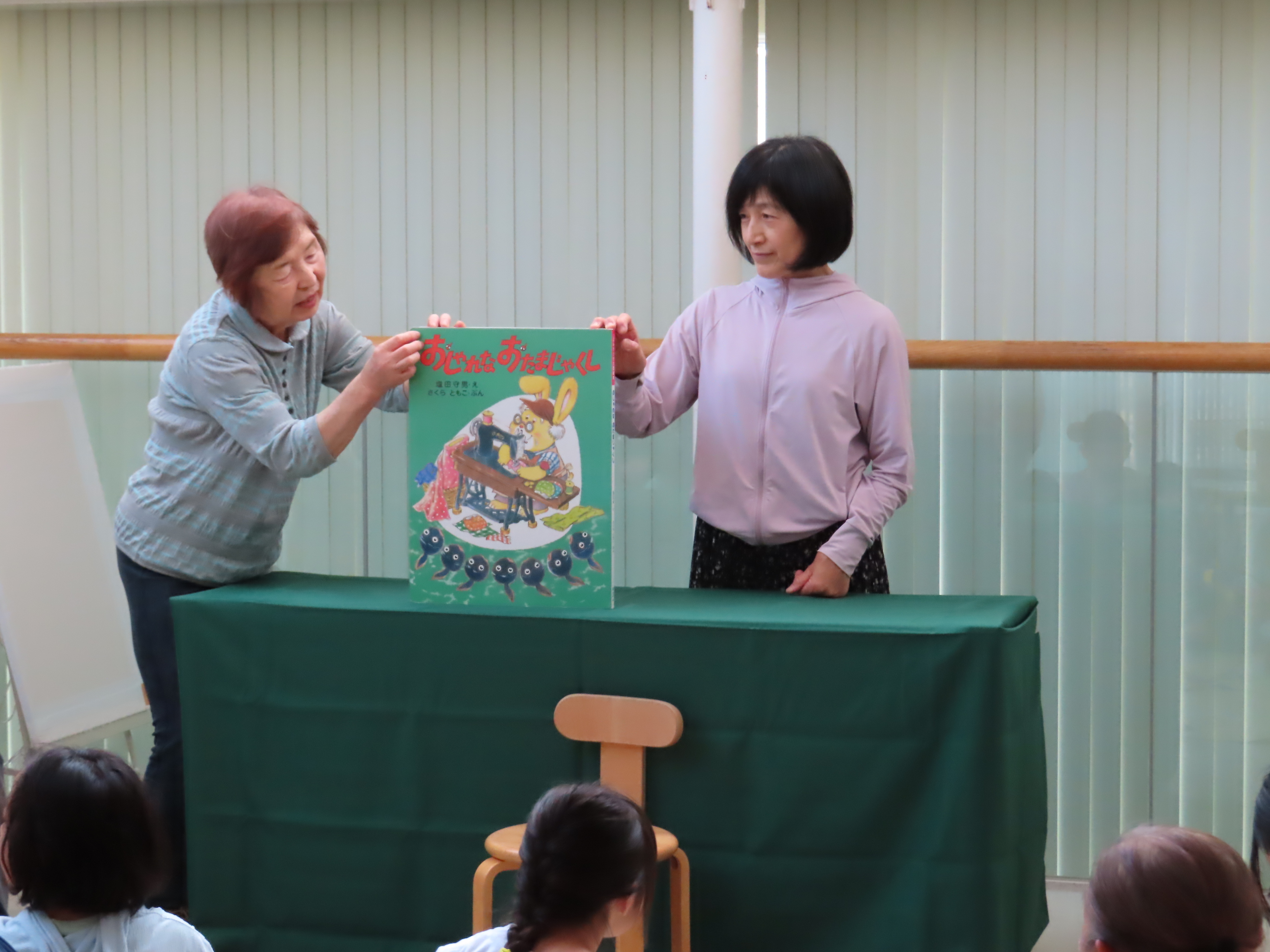 舞台前で二人の大人が絵本を子どもたちに見せている様子