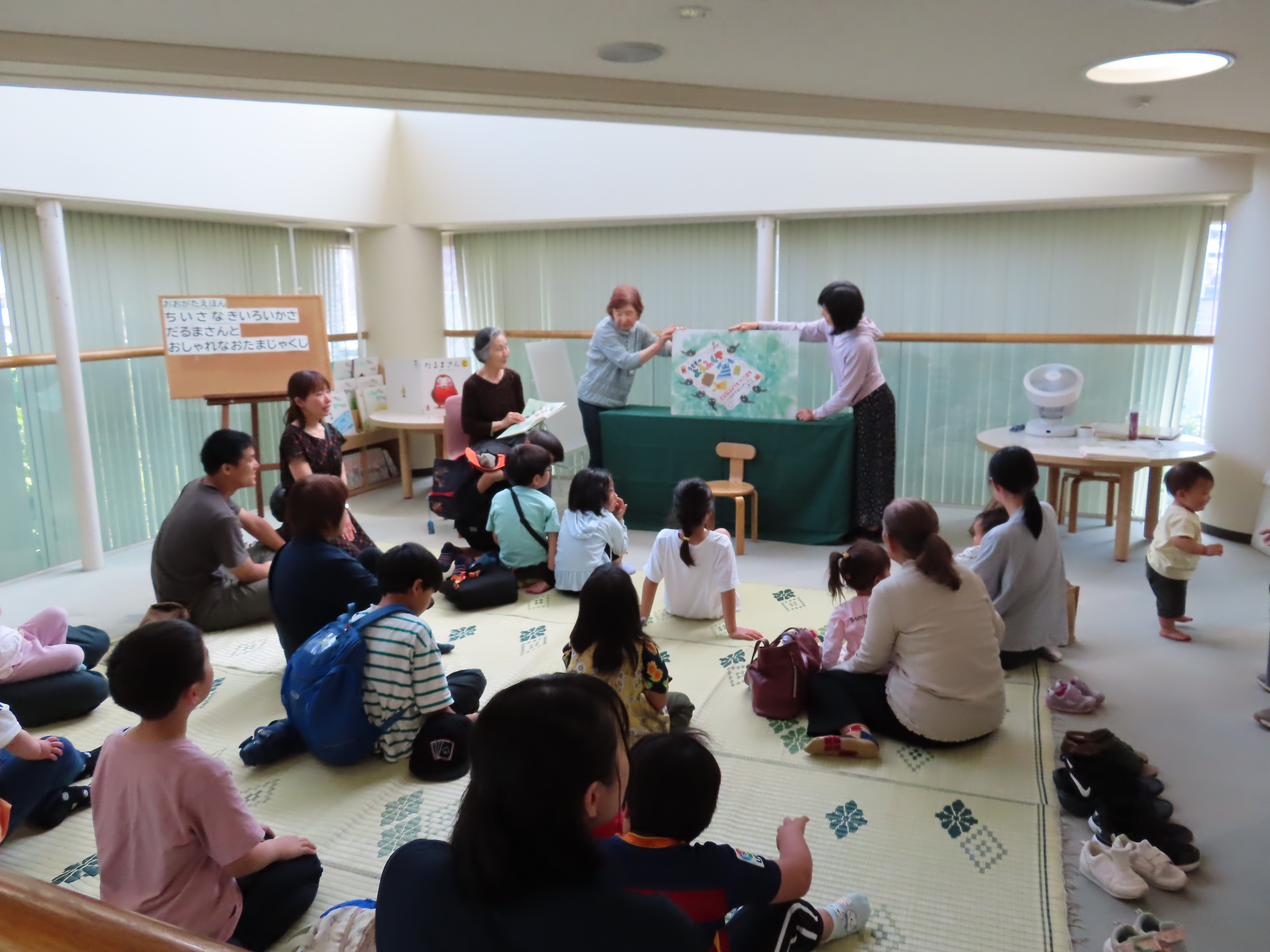 室内の広い部屋で親子や子どもたちが床に座って読み聞かせの紙芝居を見ている